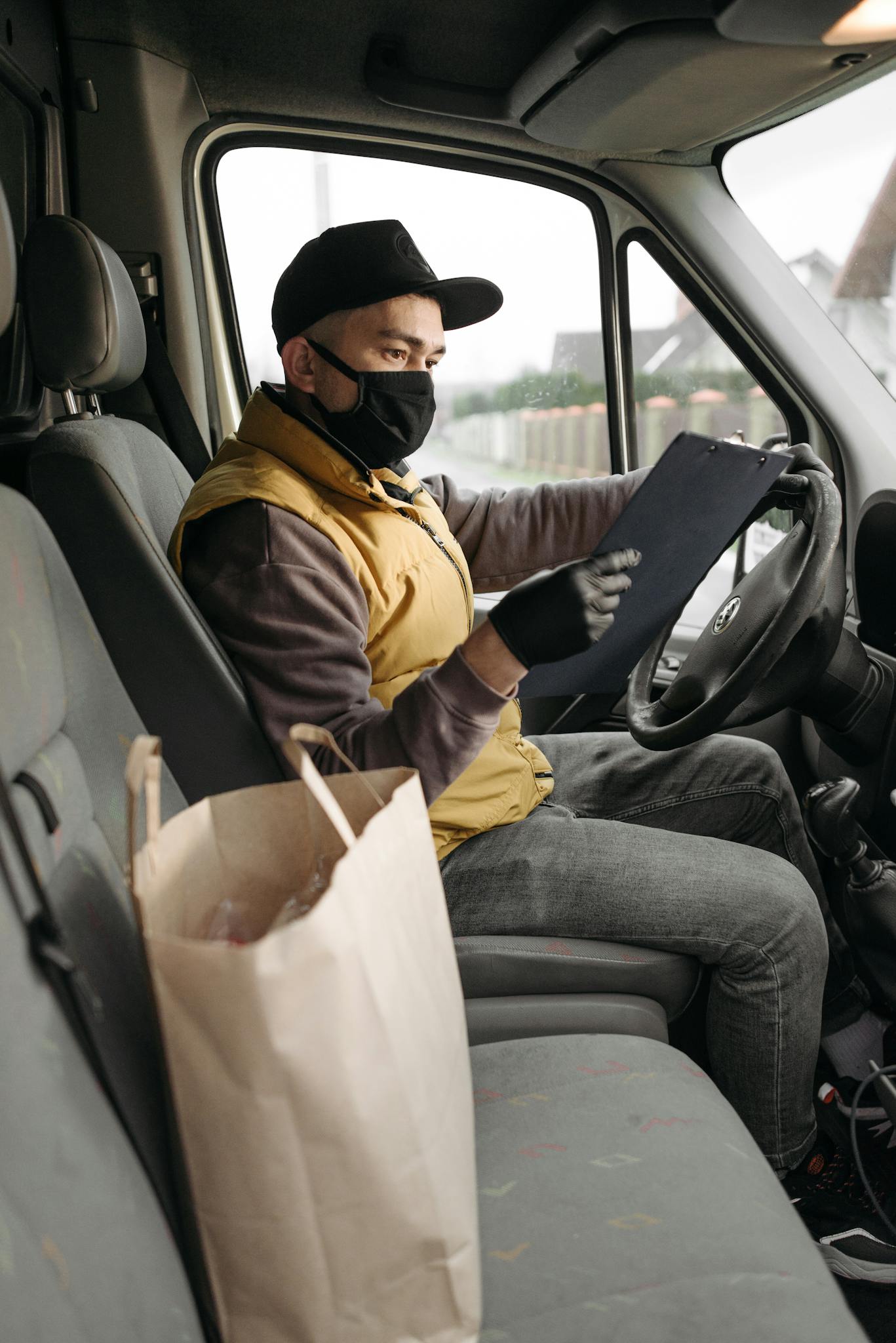 A delivery person wearing a mask holds a clipboard inside a vehicle, preparing orders for delivery.